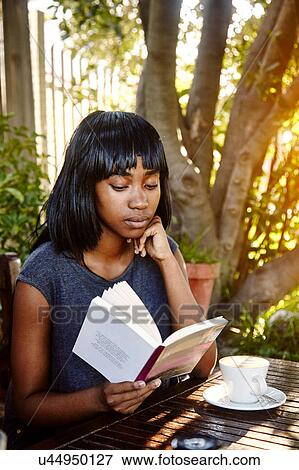 Stock Photo - Young woman sitting outdoors, reading book, coffee in front of her. Fotosearch