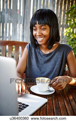 Stock Image - Young woman sitting outdoors, using laptop, drinking coffee. Fotosearch