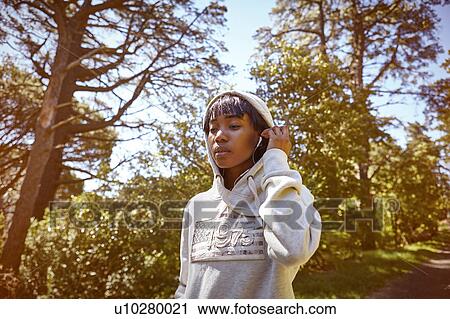 Stock Image - Young woman walking in rural setting, wearing hooded sweatshirt and earphones. Fotosearch