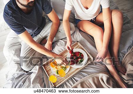 Stock Image - Couple relaxing on bed, eating strawberries, elevated view. Fotosearch