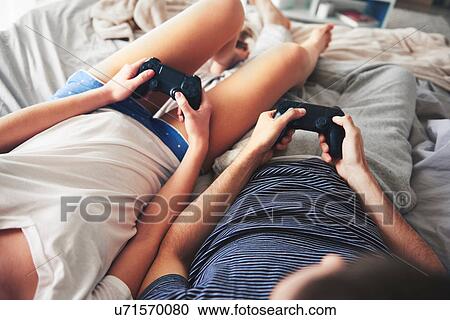 Stock Image - Couple relaxing on bed, holding computer controllers. Fotosearch