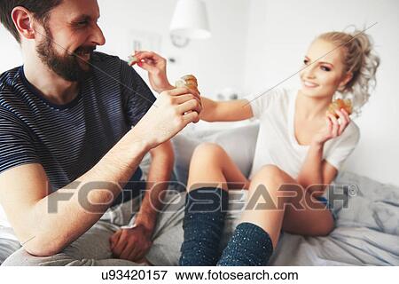 Stock Photo - Couple sitting on bed, fooling around, woman sharing pastry with man. Fotosearch