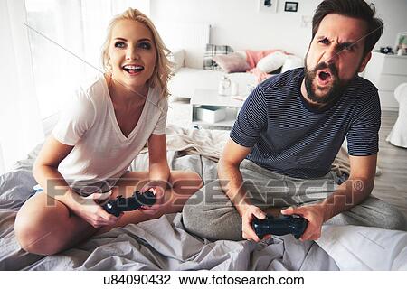 Stock Image - Couple sitting on bed, holding computer controllers, playing computer game. Fotosearch