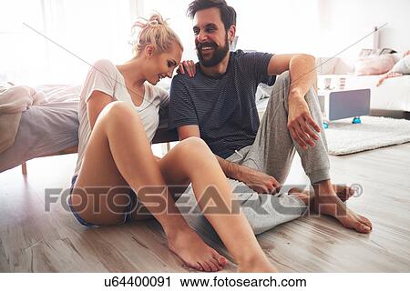 Stock Image - Couple sitting on floor beside bed, laughing. Fotosearch