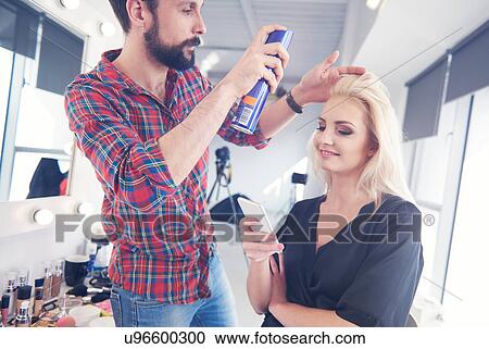 Stock Image - Male hairdresser putting hairspray on model for photo shoot. Fotosearch