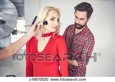 Stock Image - Male photographer and make up artist preparing female model for studio photo shoot. Fotosearch