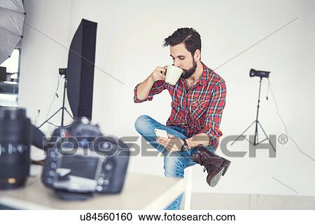 Stock Image - Male photographer drinking coffee and looking at smartphone in studio. Fotosearch