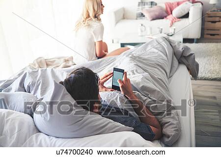 Man in bed using smartphone, woman sitting at end of bed View Large Photo Image Stock Image - Man in bed using smartphone, woman sitting at end of bed. Fotosearch