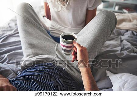 Stock Image - Man relaxing on bed, holding cup of coffee, woman sitting with her back to him. Fotosearch