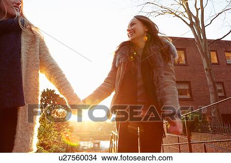 Picture - Two female friends outdoors, walking hand in hand, laughing. Fotosearch