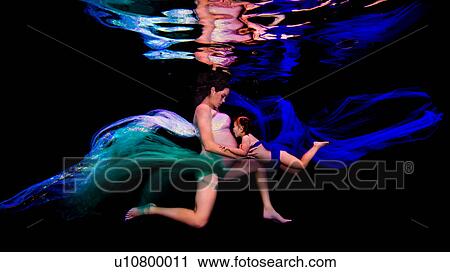 Stock Image - Underwater view of young pregnant woman and girl poised together in darkness. Fotosearch