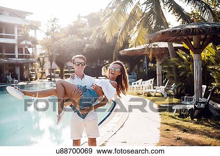 Stock Photo - Young man carrying girlfriend in arms at poolside, Koh Samui, Thailand. Fotosearch