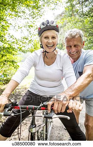 Stock Image - Mature woman on bike, Man assisting. Fotosearch