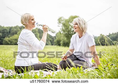 Picture - Two women sitting on grass, one drinking water. Fotosearch