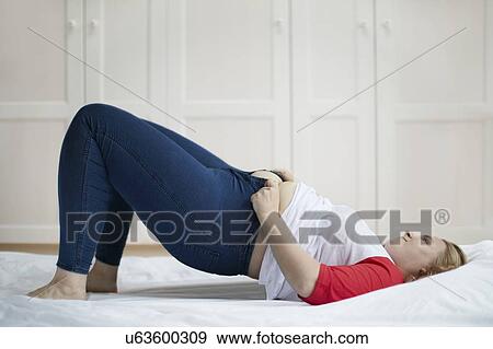 Stock Photo - Young woman lying on bed trying to button jeans. Fotosearch