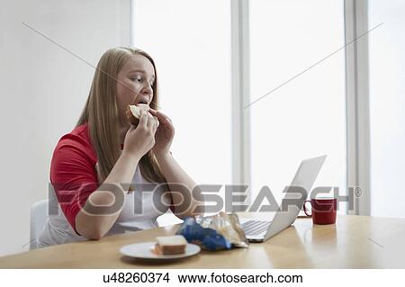 Picture - Young woman with laptop eating sandwich. Fotosearch