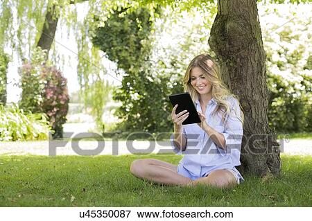 Woman sitting by tree suing digital tablet View Large Photo Image Stock Photo - Woman sitting by tree suing digital tablet. Fotosearch