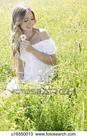 Stock Image - Woman sitting in meadow holding daisy. Fotosearch