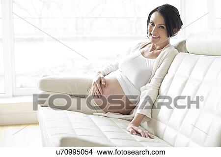 Picture - Pregnant woman on sofa touching tummy. Fotosearch