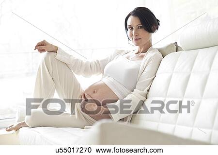 Stock Image - Pregnant woman on sofa touching tummy. Fotosearch