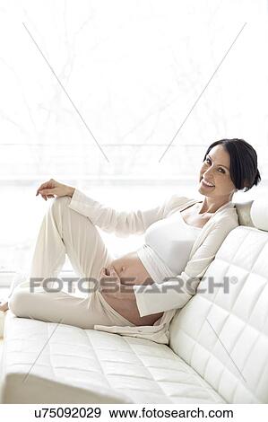 Stock Photo - Pregnant woman on sofa touching tummy. Fotosearch