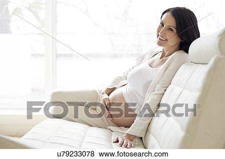 Stock Photo - Pregnant woman on sofa touching tummy. Fotosearch