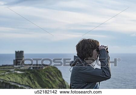 Stock Image - Ireland, Clare County, Woman looking through binoculars on Cliffs of Moher. Fotosearch