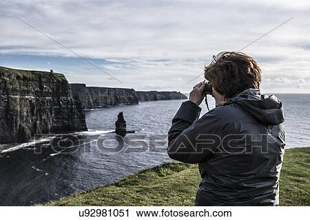 Stock Image - Ireland, Clare County, Woman looking through binoculars on Cliffs of Moher. Fotosearch
