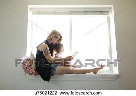 Stock Image - USA, Utah, Salt Lake City Mother sitting with daughter (12-17 months) on windowsill and reading book. Fotosearch