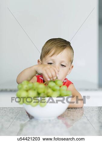 Stock Image - Boy (2-3) picking up grape from bowl. Fotosearch