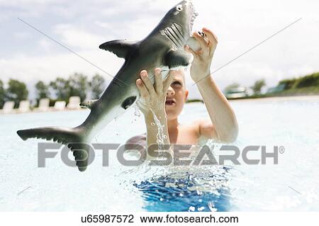 Stock Image - Boy (6-7) playing with toy shark in swimming pool. Fotosearch