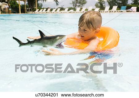 Stock Image - Boy (6-7) playing with toy shark in swimming pool. Fotosearch