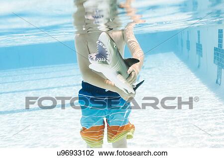 Stock Image - Boy (6-7) playing with toy shark in swimming pool. Fotosearch