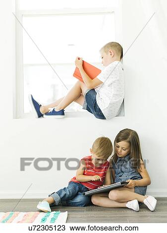 Stock Image - Boy (6-7) reading book and sister with brother (2-3, 6-7) using digital tablet. Fotosearch