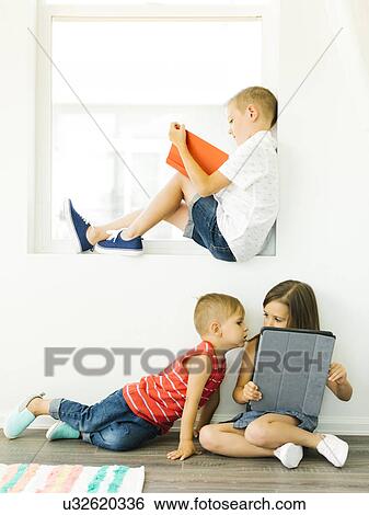 Stock Photograph - Boy (6-7) reading book and sister with brother (2-3, 6-7) using digital tablet. Fotosearch