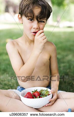 Stock Photo - Boy (6-7) sitting on grass and eating strawberry in park. Fotosearch