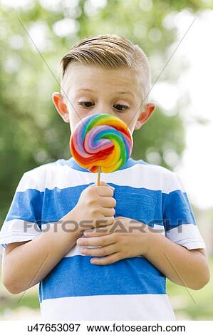 Boy (6-7) squinting and holding big, colorful lollipop View Large Photo Image Stock Photo - Boy (6-7) squinting and holding big, colorful lollipop. Fotosearch