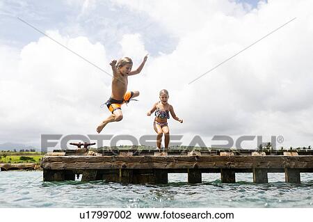 Stock Image - Boy (8-9) and girl (6-7) jumping into sea from jetty. Fotosearch