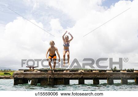 Stock Photograph - Boy (8-9) and girl (6-7) jumping into sea from jetty. Fotosearch
