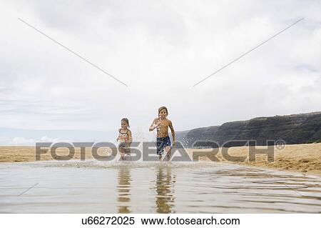 Stock Photography - Boy (8-9) and girl (6-7) running through sea. Fotosearch