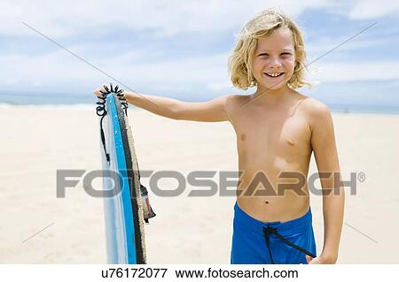 Stock Photo - Boy (8-9) standing on beach and holding surfboard. Fotosearch