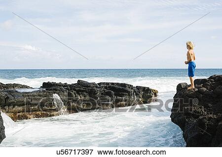 Stock Photo - Boy (8-9) standing on rock against blue sky. Fotosearch