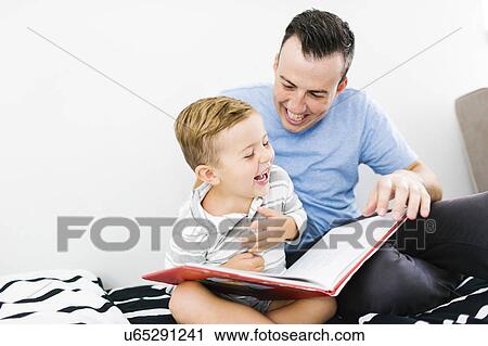 Stock Image - Father and son (4-5) sitting on bed and holding book. Fotosearch