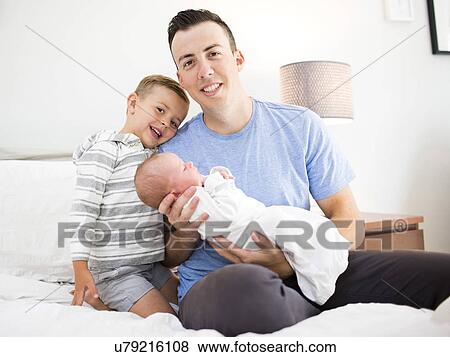Stock Photo - Father carrying daughter (0-1 months) and sitting on bed with son (4-5). Fotosearch