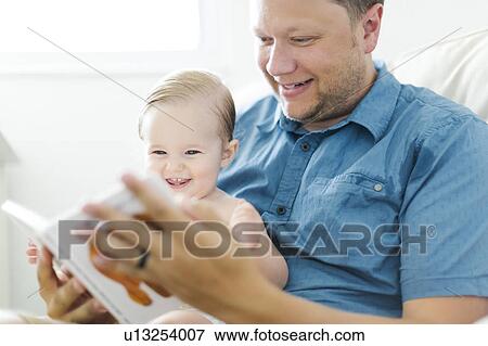 Father with baby girl (12-17 months) sitting in living room and reading book View Large Photo Image Stock Photo - Father with baby girl (12-17 months) sitting in living room and reading book. Fotosearch