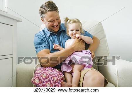Father with baby girl (12-17 months) sitting in living room View Large Photo Image Stock Photo - Father with baby girl (12-17 months) sitting in living room. Fotosearch