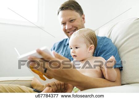 Stock Photo - Father with baby girl (12-17 months) sitting in living room and reading book. Fotosearch