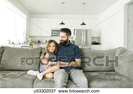 Stock Photo - Father with daughter (6-7) sitting on sofa in living room. Fotosearch