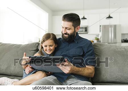 Stock Image - Father with daughter (6-7) using digital tablet in living room. Fotosearch