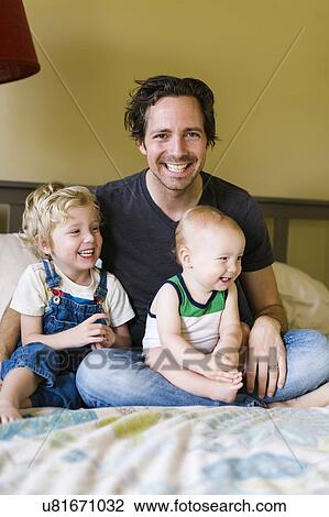 Stock Image - Father with sons (12-17 months, 4-5) sitting on bed. Fotosearch